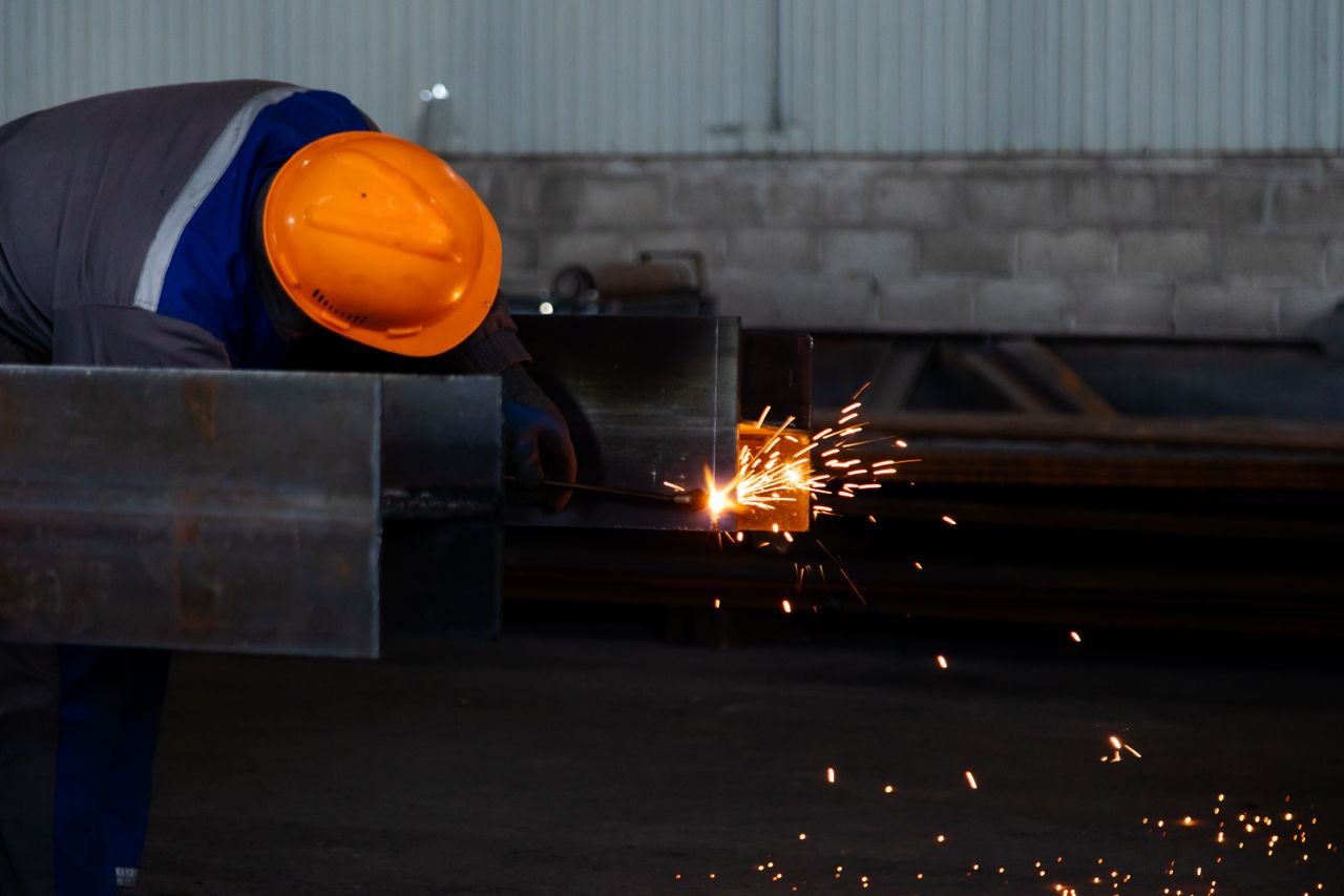 our-story-01 Worker wearing safety gear welds metal beam, emitting sparks in a factory setting.